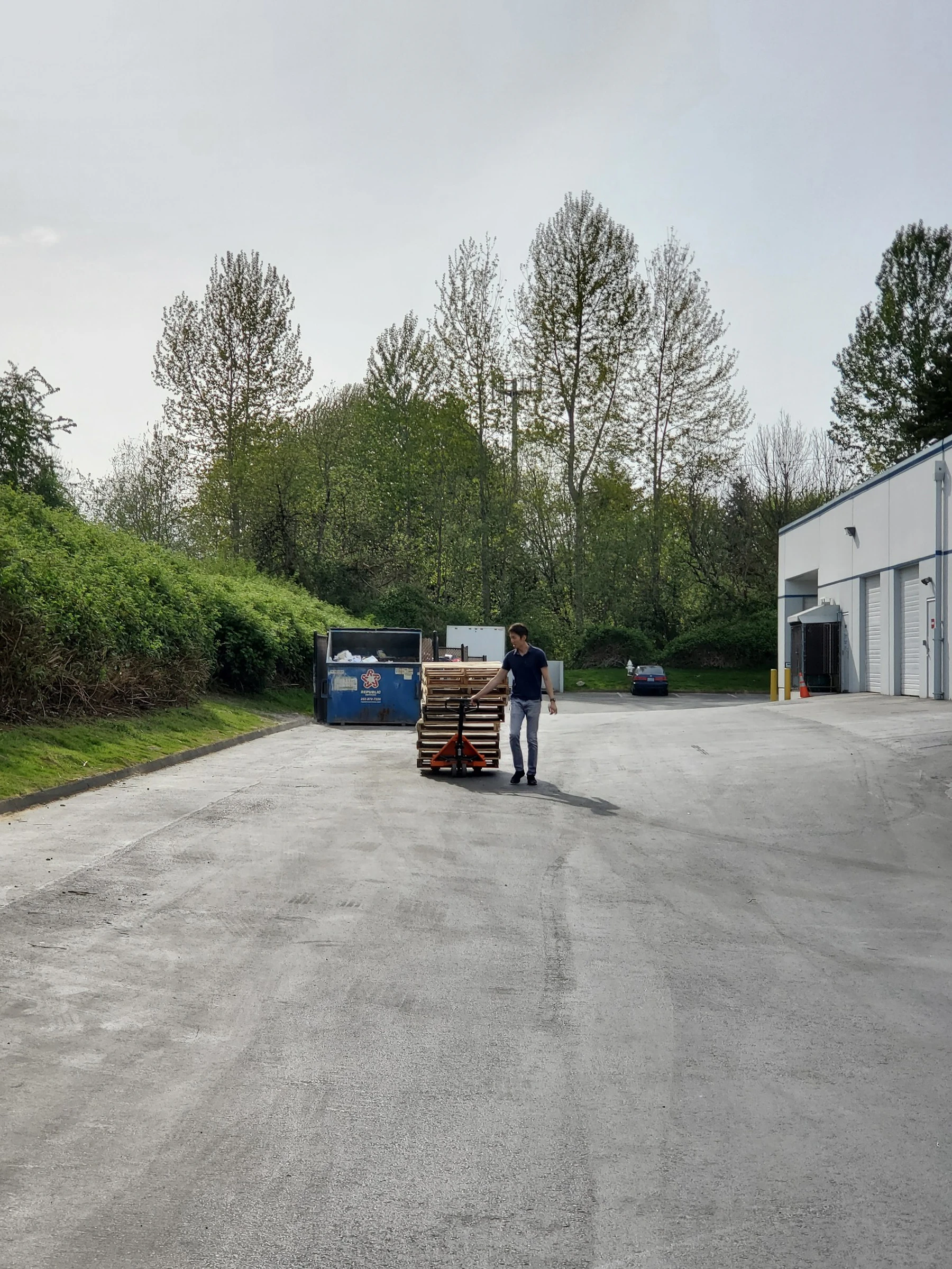 Worker moving stacked wooden pallets with a manual pallet jack outside an industrial building